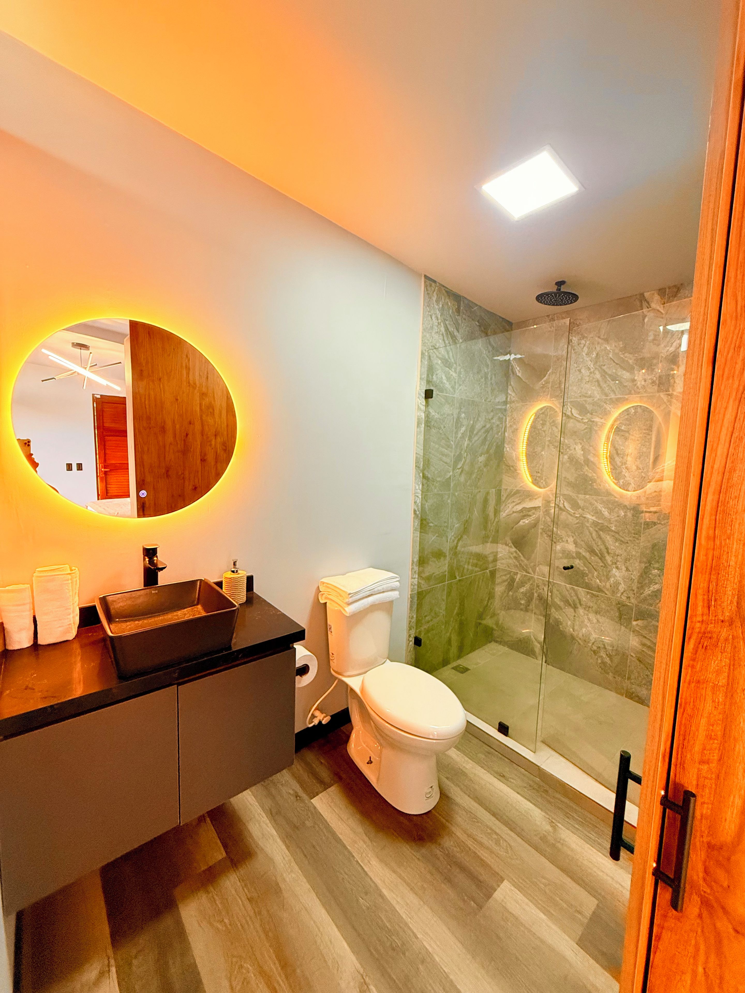 A modern bathroom with a wooden vanity, a glass shower enclosure, and a round mirror with LED lighting.