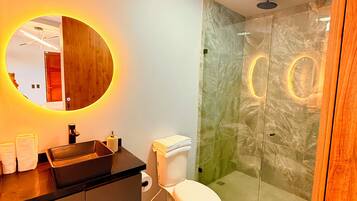 A modern bathroom with a wooden vanity, a glass shower enclosure, and a round mirror with LED lighting.