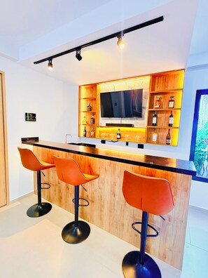 A modern kitchen with a bar area, wooden bar stools, a television, and shelves with bottles.
