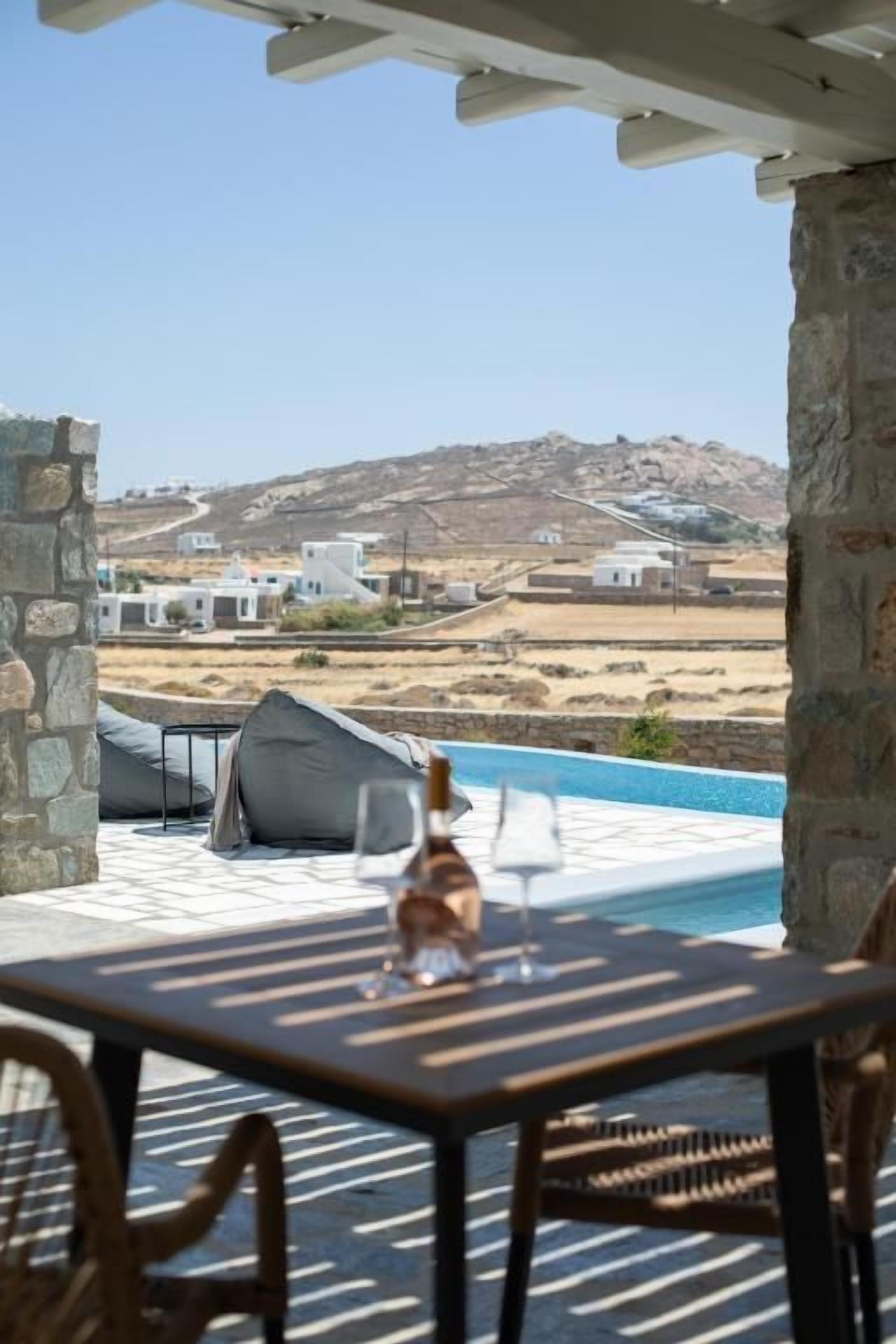 Un espace aménagé au bord de la piscine, avec une table et des chaises, une bouteille de vin, et une vue sur des bâtiments blancs et des collines au loin.
