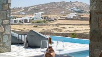 Zona junto a la piscina con una mesa y sillas, una botella de vino, y vistas a edificios blancos y colinas a lo lejos.