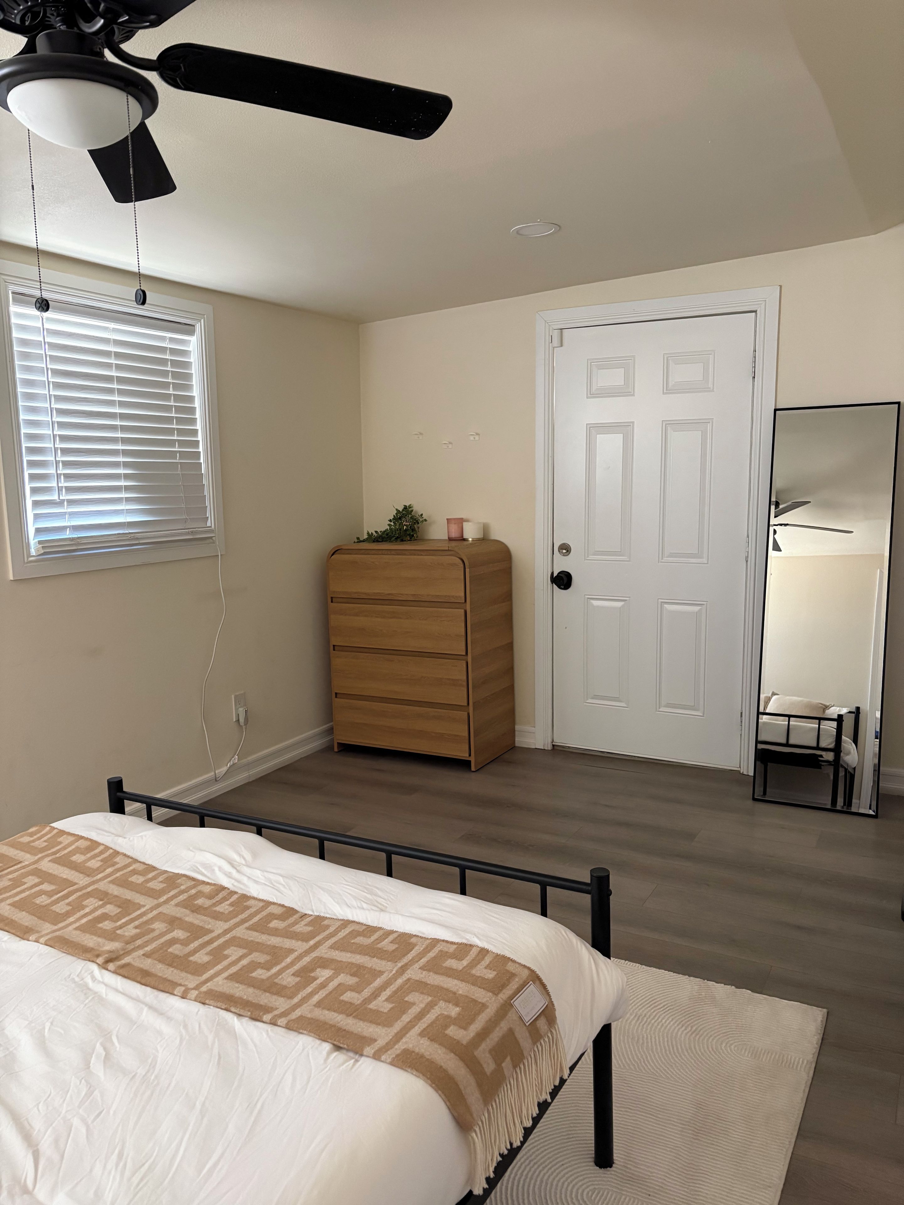 Une chambre à coucher comprenant un lit, une commode, un ventilateur de plafond, une fenêtre avec des stores, une porte et un miroir.