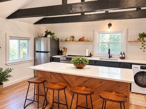 A kitchen with a central island, wooden stools, a refrigerator, a washing machine, and wooden beams on the ceiling. - Maple Cabin w Sauna, Indoor Hot Tub, + Firepit (Greentown)