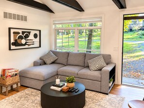 A modern living room with a grey sectional sofa, a round black coffee table, and a large window with a view of greenery. - Maple Cabin w Sauna, Indoor Hot Tub, + Firepit (Greentown)