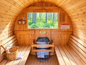 A sauna room with wooden walls, a large window with a forest view, a sauna heater, and a wooden bucket. - Maple Cabin w Sauna, Indoor Hot Tub, + Firepit (Greentown)