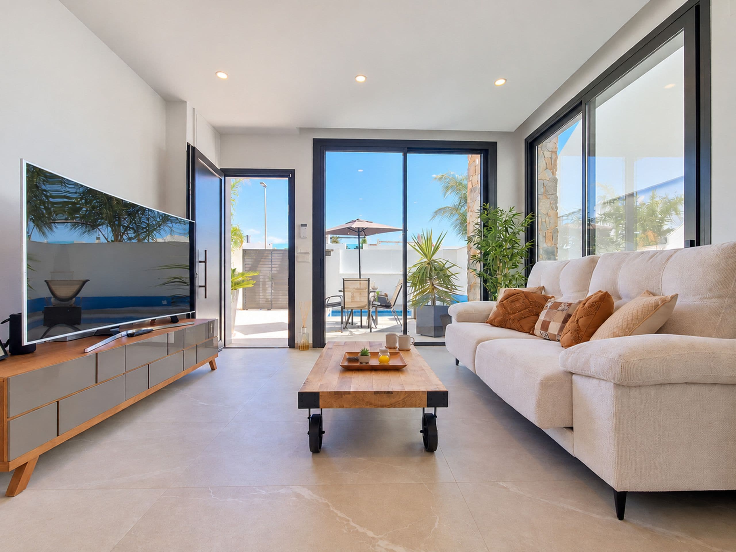 A modern living room with a large flat-screen TV, a sofa, a coffee table, and a view of the outdoors through large sliding doors.