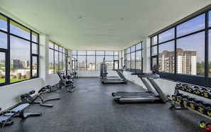 Ein Fitnessstudio mit Laufbändern, Kraftgeräten und Kurzhanteln, mit großen Fenstern und Blick auf die Stadt.