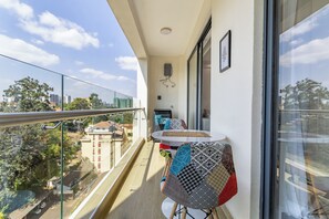 Een balkon met een glazen hekwerk, een klein tafeltje met een kussen, en uitzicht op de stad.