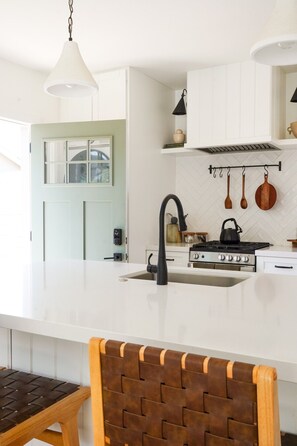 A modern kitchen with a white countertop, a black faucet, and a stove. There is a wooden chair with a woven backrest. - Modern Family Home & Guest House - Sleeps 9 (Ojai)