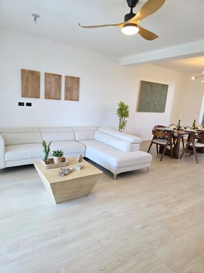A modern living room with a sectional sofa, a dining table set for four, and a ceiling fan. - Luxury Penthouse | 2BR | 2 Terraces | Estrrlla Garden Condos (Dominicus)