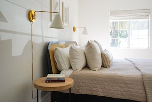 Une chambre à coucher avec un lit, des oreillers, une table de chevet avec des livres et des lampes fixées au mur.