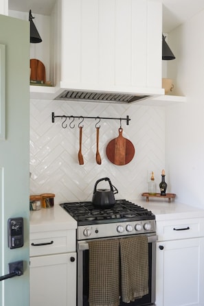 A kitchen with a gas stove, white cabinets, a herringbone-patterned backsplash, and hanging utensils.