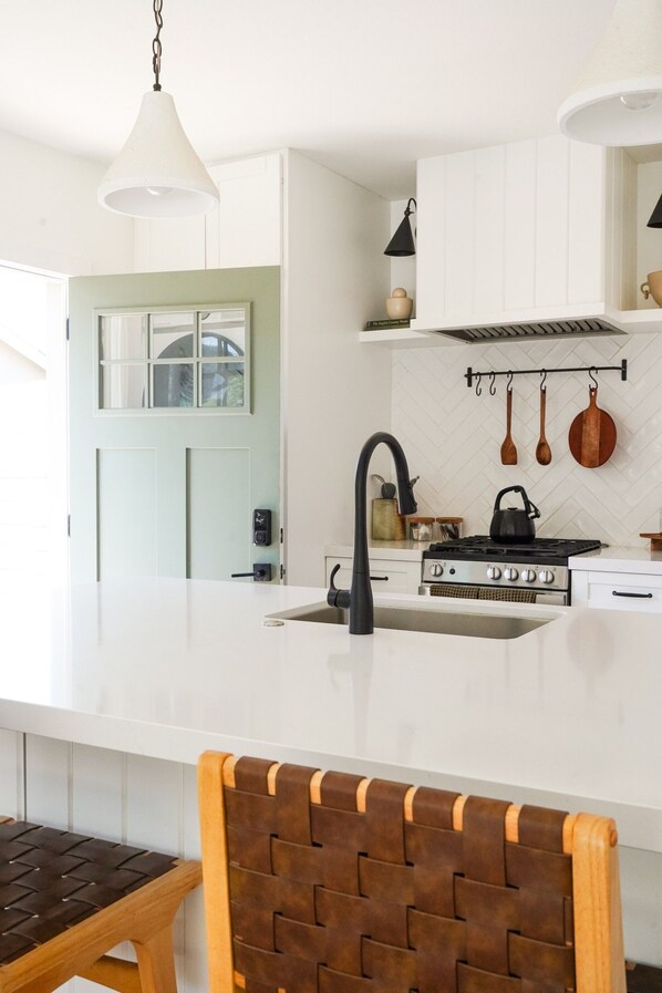 A modern kitchen with a white countertop, a black faucet, and a stove. There is a wooden chair with a woven backrest.