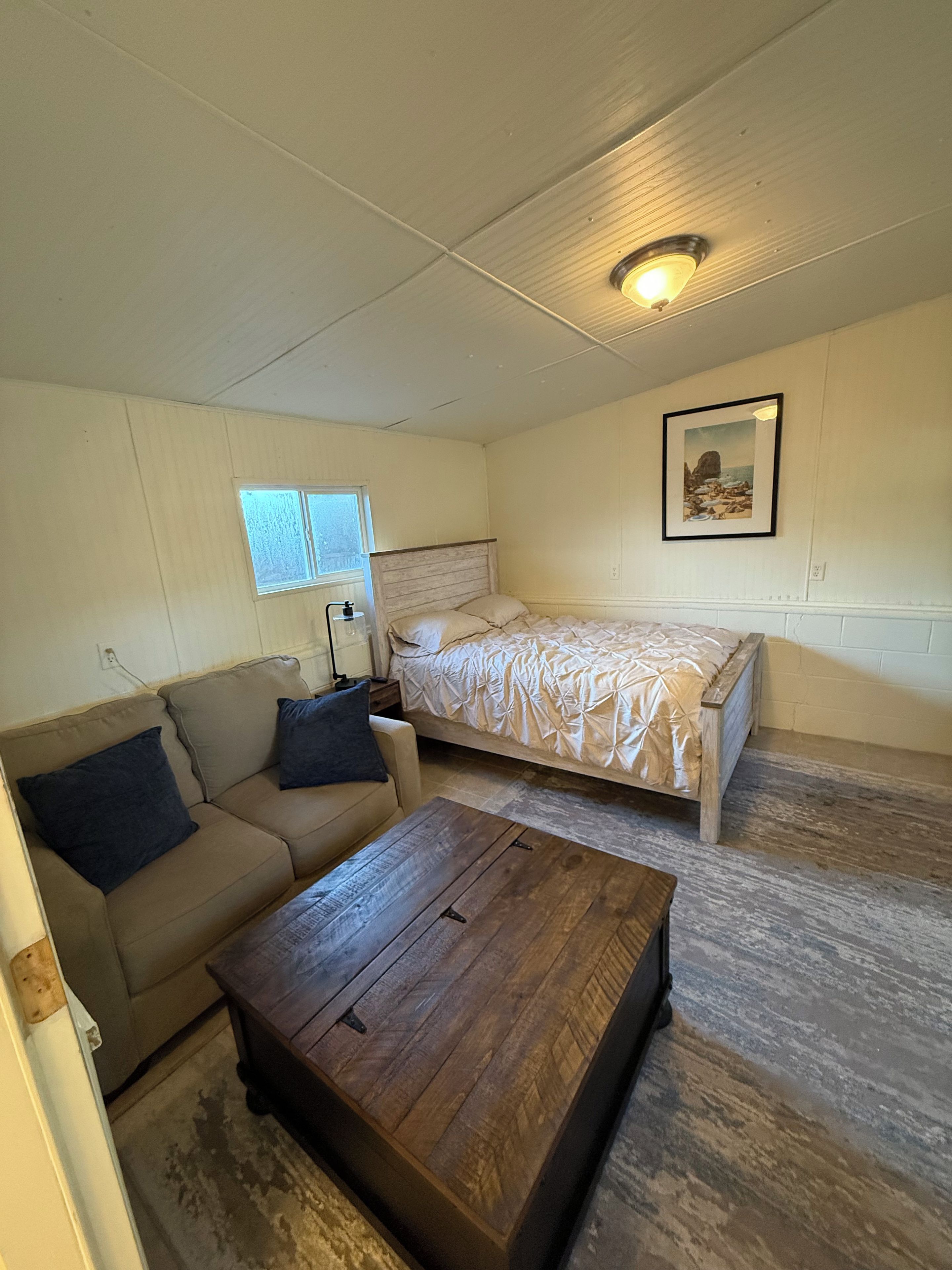 A bedroom with a bed, a sofa, a wooden coffee table, a window, and a wall-mounted light fixture.