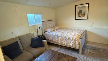 A bedroom with a bed, a sofa, a wooden coffee table, a window, and a wall-mounted light fixture.