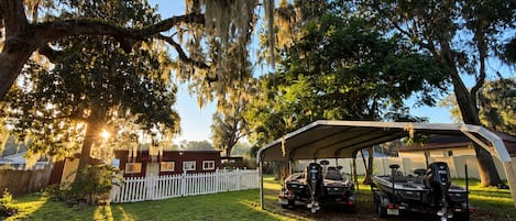 A covered outdoor area with a vehicle and a boat, surrounded by trees and a white fence.