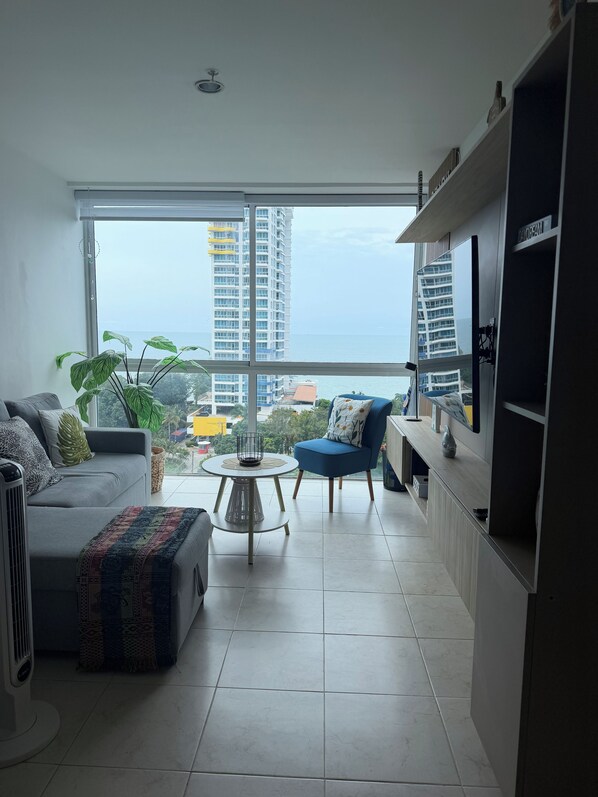 A modern living room with a sofa, armchair, and a small coffee table. A large window offers a view of the city and the sea.