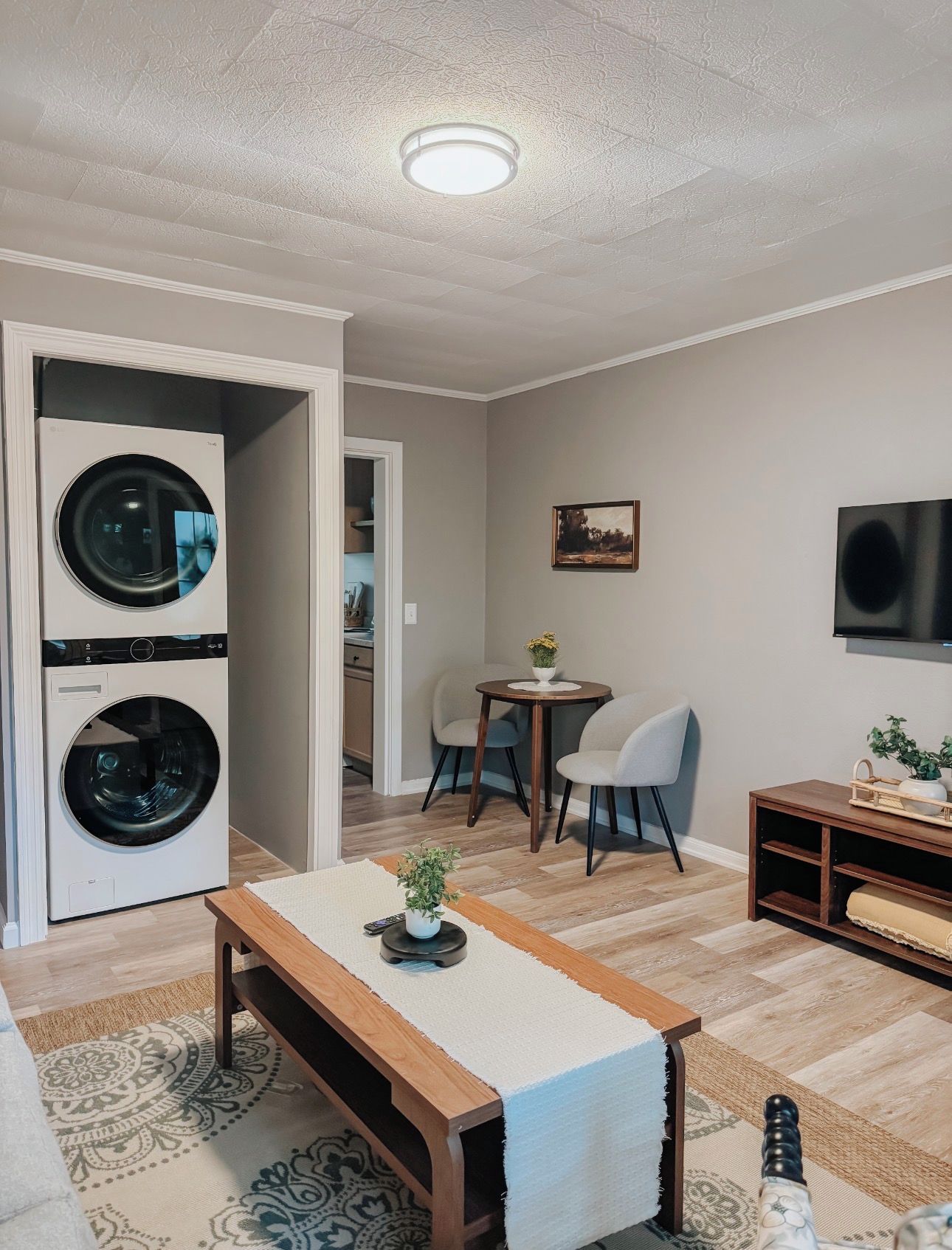 Un salon comprenant un lave-linge, une table à manger avec des chaises, un téléviseur fixé au mur et un tapis à motifs.