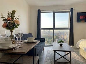 Uma sala de estar moderna com uma mesa de jantar posta para quatro pessoas, um sofá, uma pequena mesa com um bonsai e uma janela grande com vista para a cidade.