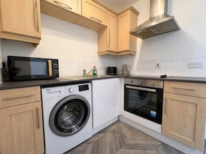 A compact kitchen with a washing machine, microwave, oven, and wooden cabinets. - AO Arena • Parking • Victoria Station (Manchester)