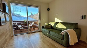 Ein Zimmer mit einem grünen Sofa, Holzboden und einer Schiebetür zum Balkon. Im Hintergrund ist eine Seilbahn zu sehen.