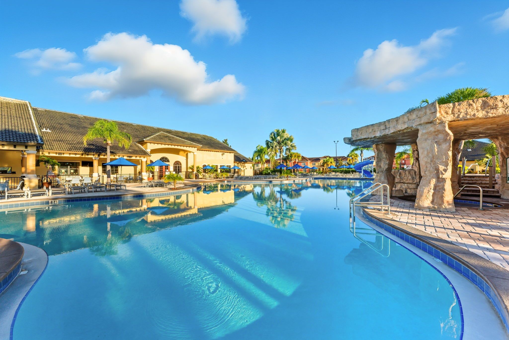 A swimming pool with a clear blue water surface, surrounded by lounge chairs and umbrellas, with buildings in the background.