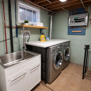 A laundry room with a stainless steel sink, two front-loading washing machines, a shelf with cleaning supplies, and a wall-mounted electrical panel. - Modern 4BR Home | Subway | Parking | Toronto (Toronto)