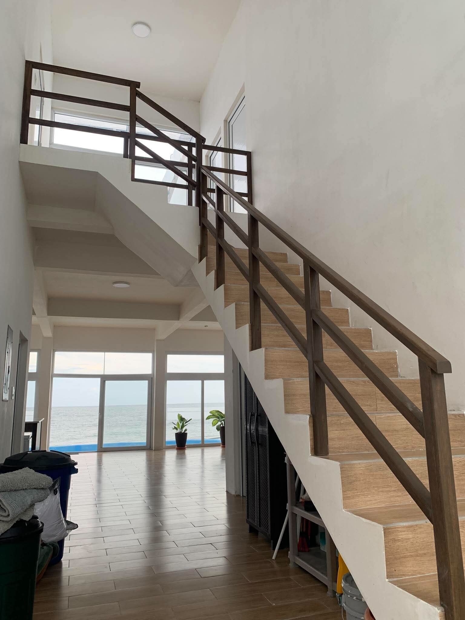 Un interior espacioso con una escalera de madera que lleva a un nivel superior, grandes ventanales con vistas al mar y un diseño moderno y cuidado.