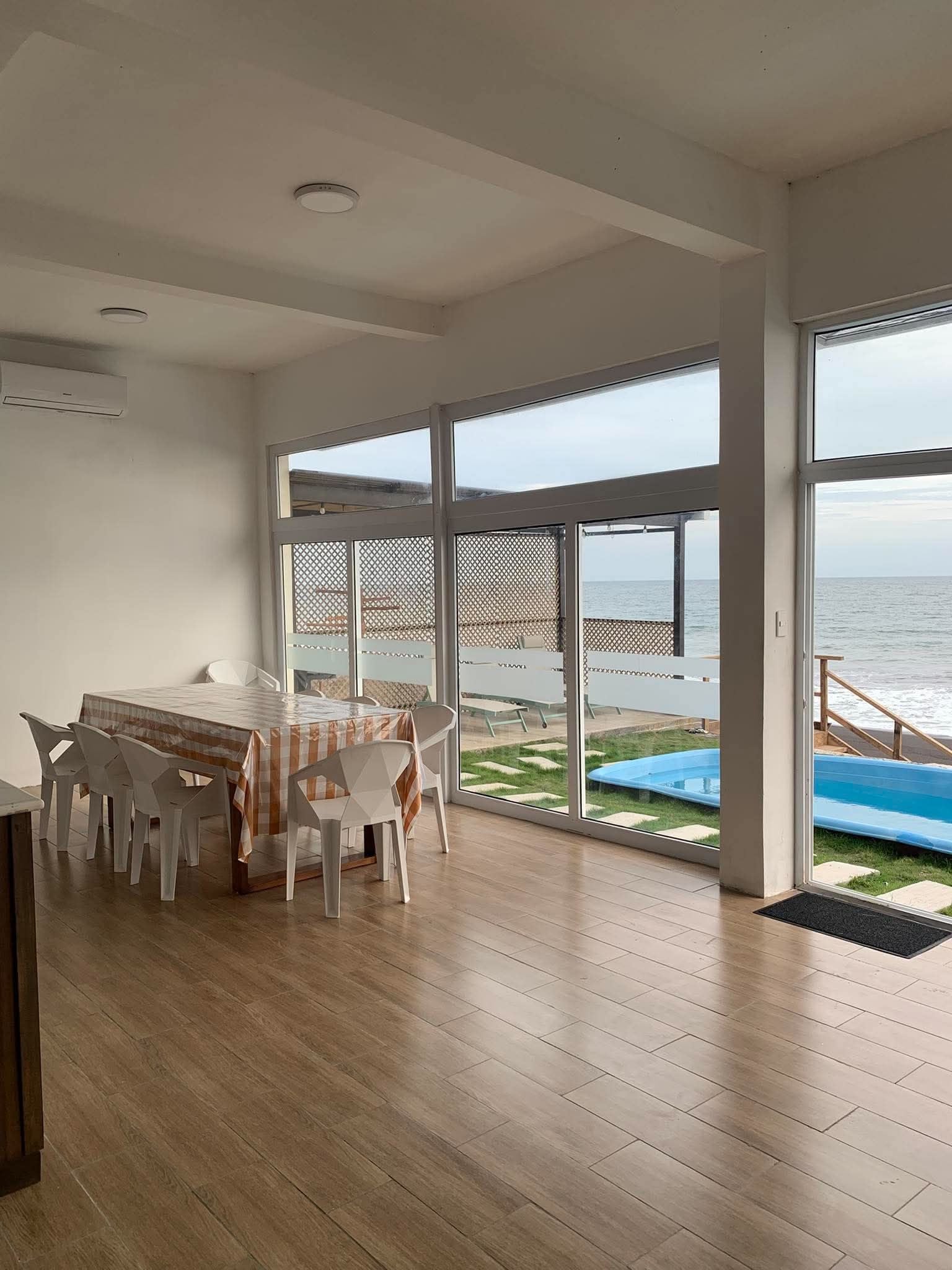 Une pièce spacieuse dotée d’un plancher en bois, d’une table à manger avec des chaises, et de grandes fenêtres offrant une vue sur la mer et une piscine.