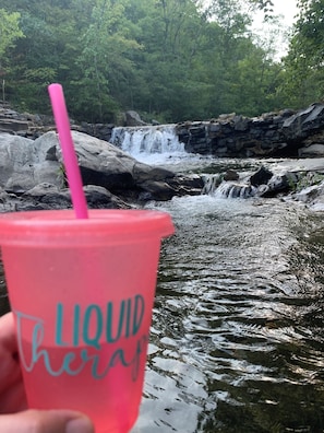 A person holding a pink cup with 'LIQUID heraf' text, in front of a waterfall and rocky terrain.