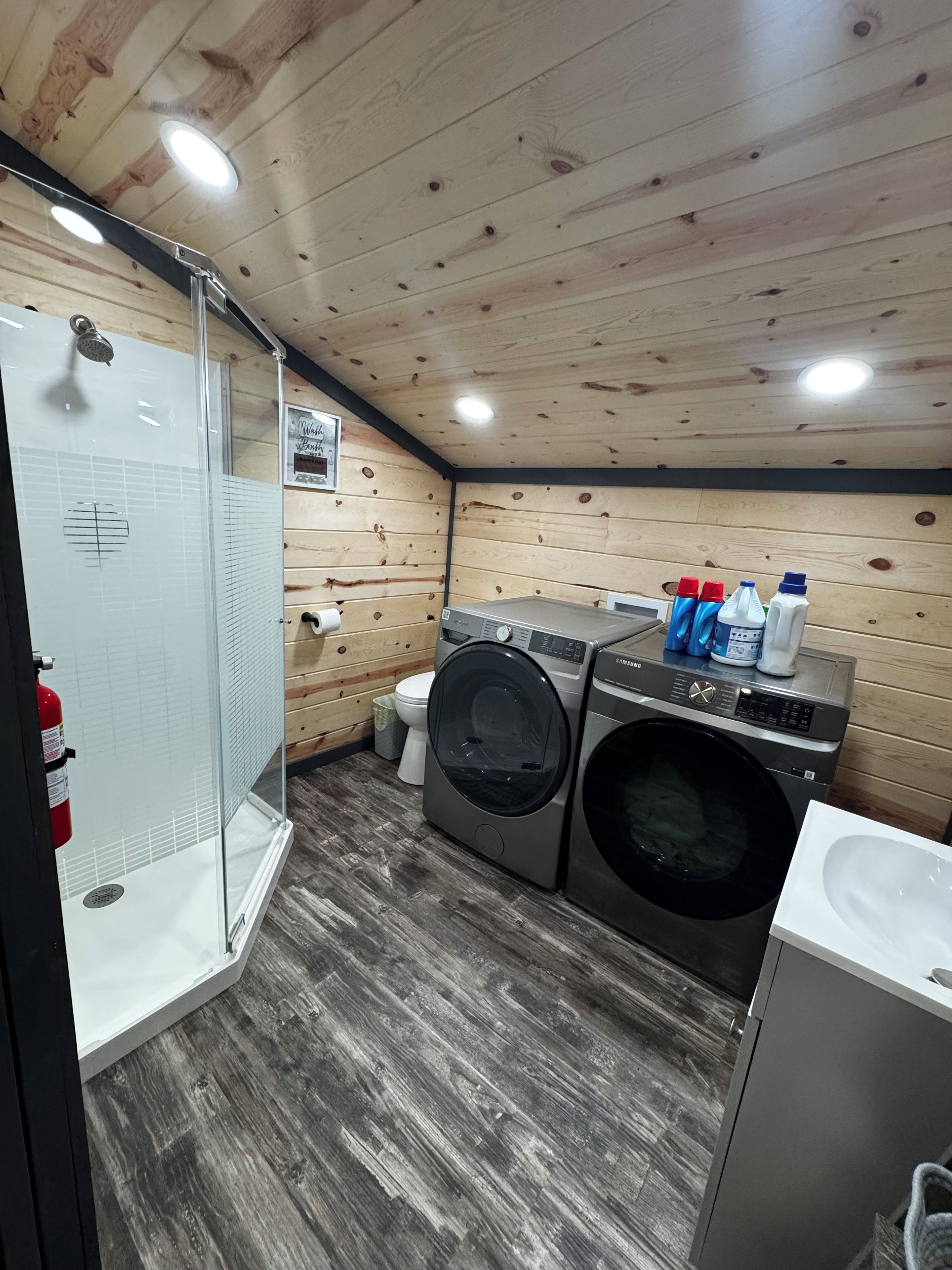 Une petite buanderie comprenant un lave-linge, un sèche-linge et une douche fermée par une paroi en verre.