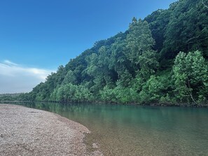 Orilla de río con agua cristalina y una ladera boscosa.