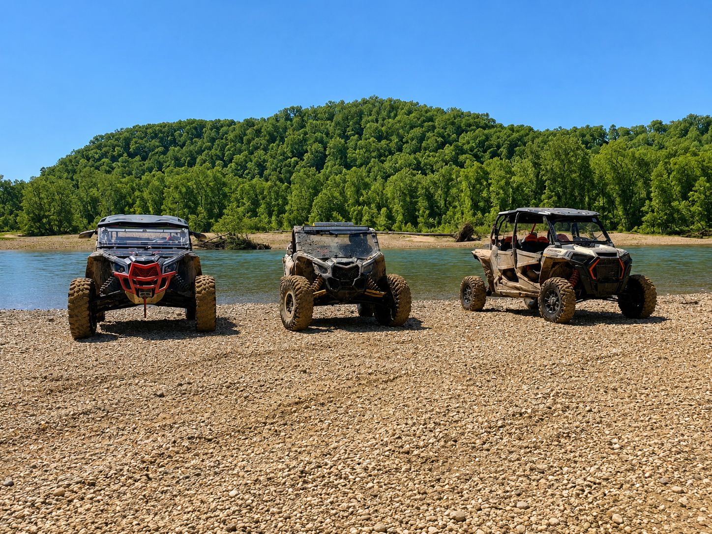 Three off-road vehicles parked on a pebbled beach with a forested hill in the background.