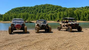 Three off-road vehicles parked on a pebbled beach with a forested hill in the background.
