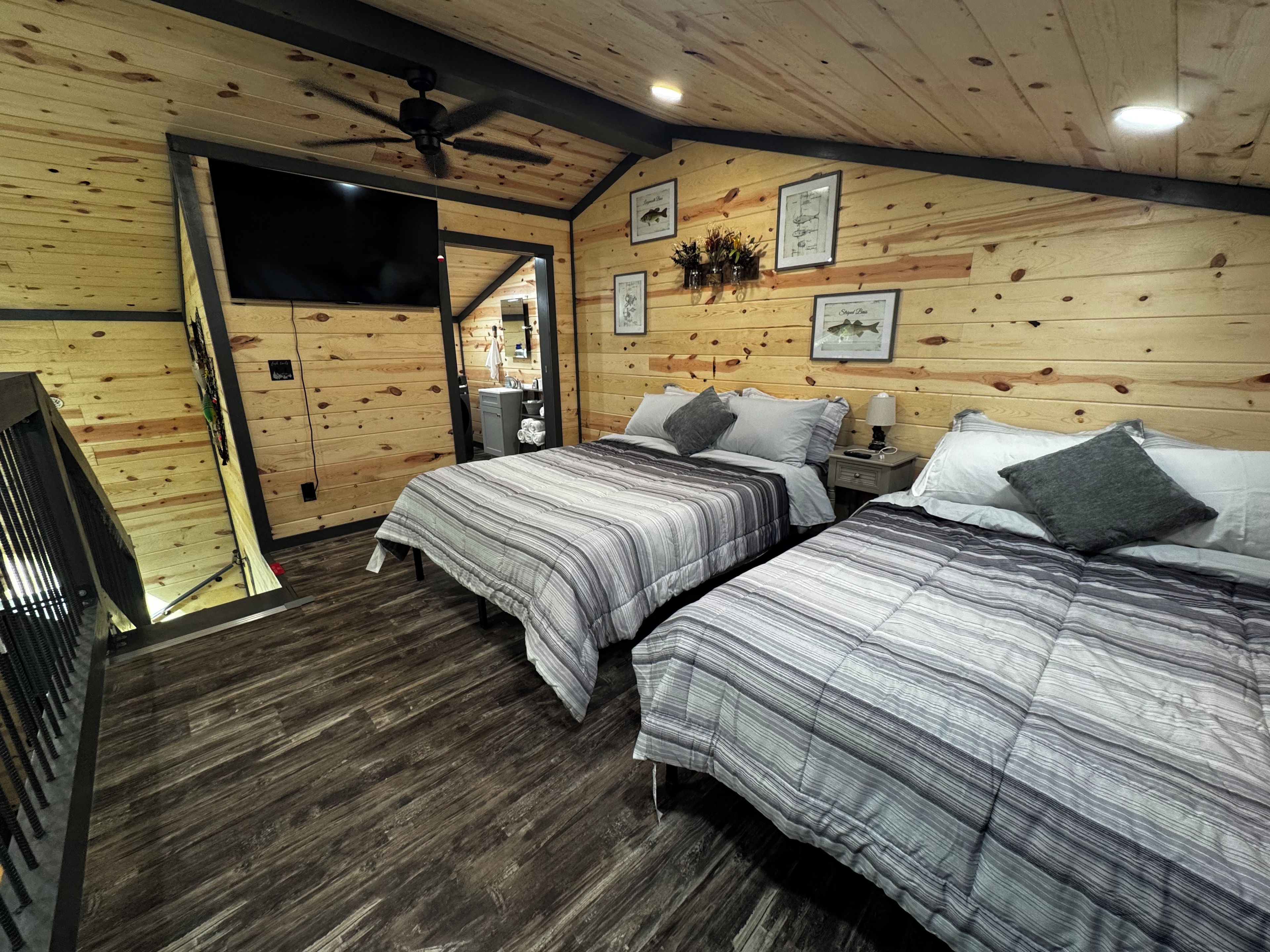 Une chambre avec deux lits, des murs en bois, un ventilateur de plafond, une télévision et des tableaux encadrés.