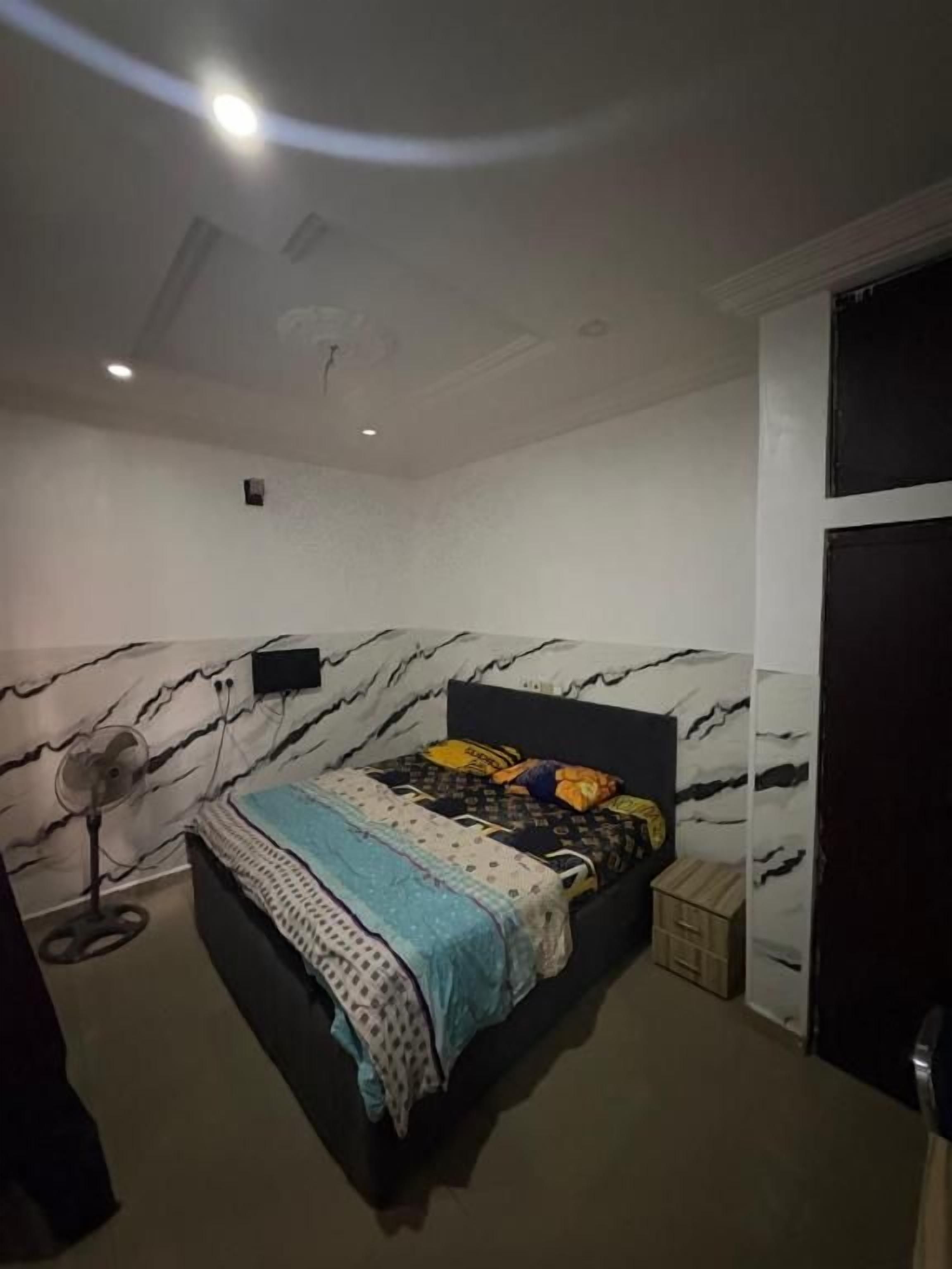 Un dormitorio con una cama, un ventilador de pie y un pequeño cofre de madera.