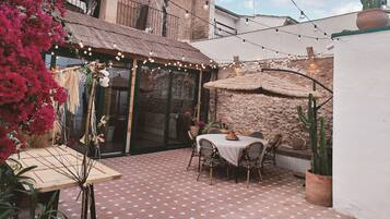 Une terrasse avec une table ronde et des chaises, entourée de plantes en pot et agrémentée d’un parasol décoratif.