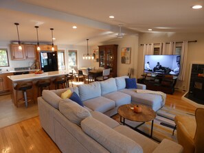 A modern living room with a large sectional sofa, a dining area with a kitchen in the background, and a flat-screen TV. - Barefoot Shores at Torch Lake (Rapid City)
