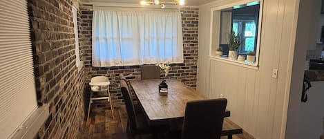 A dining area with a wooden table, chairs, a window with curtains, and a brick wall.