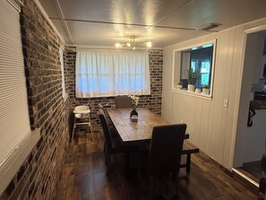 Un coin repas avec une table en bois, des chaises, une fenêtre avec des rideaux et un mur en briques.