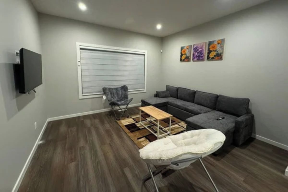 A living room with a grey sofa, a flat-screen TV mounted on the wall, a folding chair, and a small wooden coffee table.