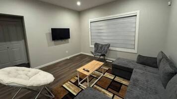 A living room with a grey sofa, a white chair, a small wooden table, and a television mounted on the wall.