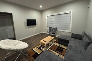 A living room with a grey sofa, a white chair, a small wooden table, and a television mounted on the wall. - 3BHK Close to Airport with Backyard (Edmonton)