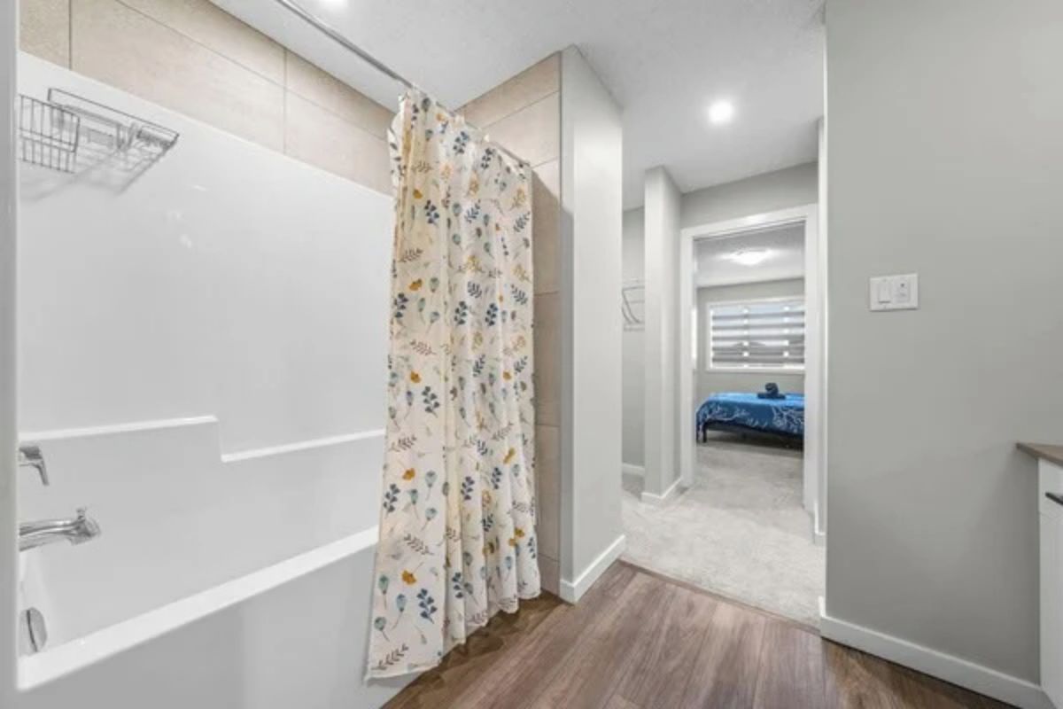 A bathroom with a shower curtain, a bathtub, and a toilet. There is a hallway leading to a bedroom with a bed.