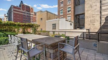 An outdoor patio with a stone table and metal chairs, surrounded by a brick wall and greenery.