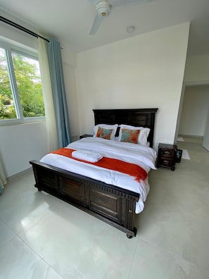 Quarto com cama grande, ventilador de teto, janela com cortinas e mesa de cabeceira.