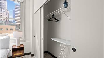 Een hotelkamer met een bed, nachtkastje, radiator, raam met uitzicht op gebouwen, een wandplank voor een strijkplank en een kledingrek.