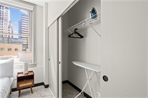 A hotel room with a bed, a nightstand, a radiator, a window with a view of buildings, a wall-mounted ironing board, and a hanging clothes rack. - Central Flat: Free Attached Parking | Rooftop |Gym (Kansas City)