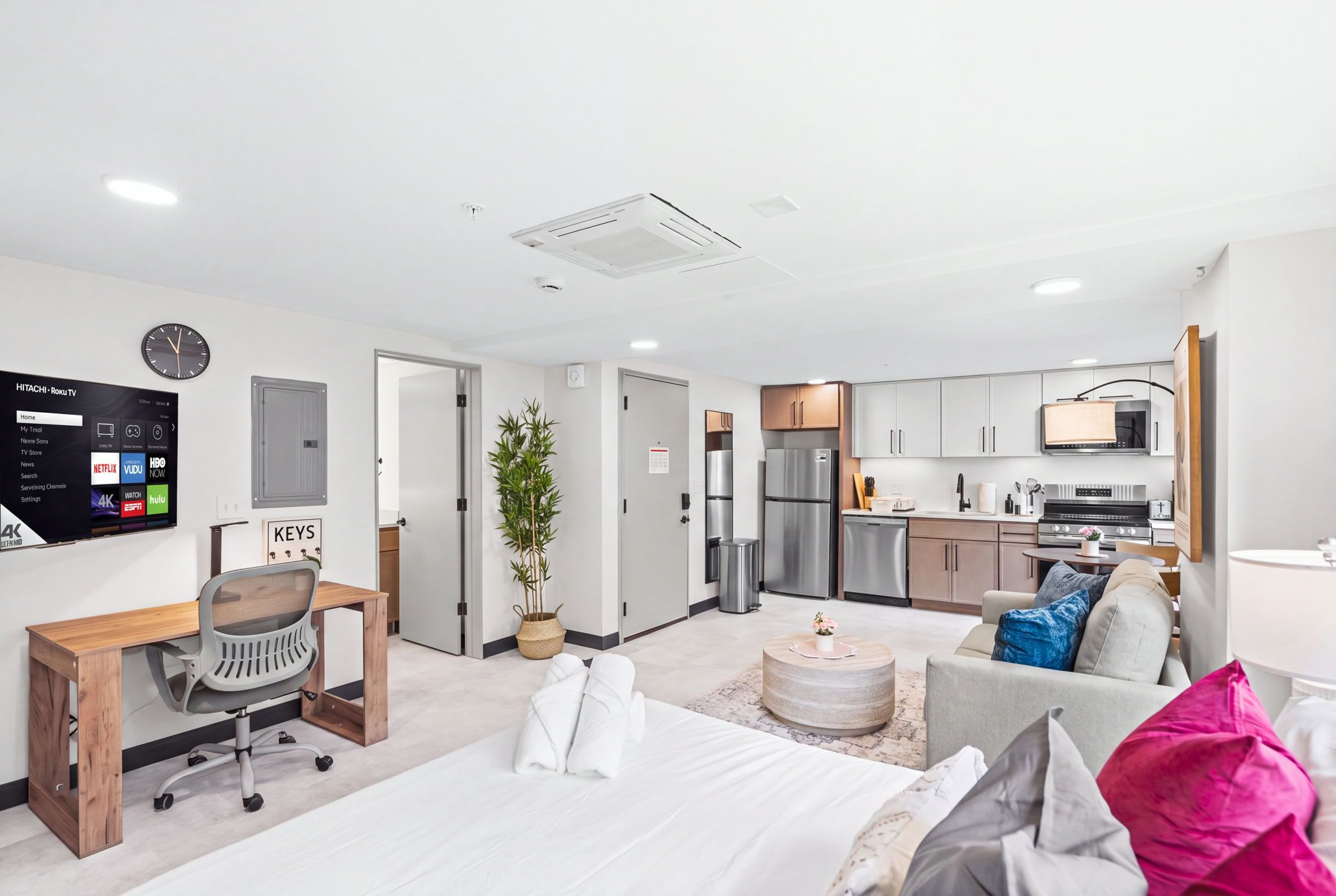 A modern hotel room with a bed, a desk, a chair, a TV, and a kitchenette.