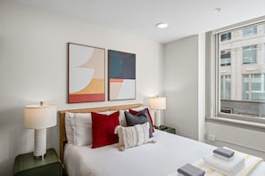 Ett hotellrum med en säng, två sänglampor, ett fönster med utsikt över en byggnad och två abstrakta målningar på väggen.